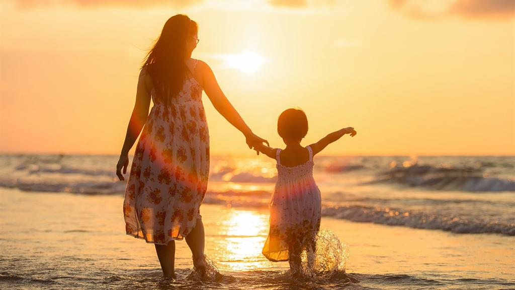 Eine Mutter und ein kleines Mädchen halten sich an der Hand und gehen im flachen Wasser, im Gegenlicht bei Sonnenuntergang